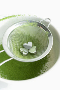 Overhead view of three heart-shaped stainless steel charms resting in a mesh matcha sifter above a bowl of sifted matcha