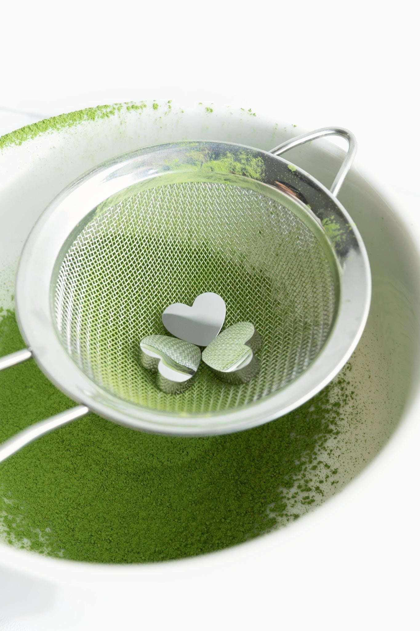 Overhead view of three heart-shaped stainless steel charms resting in a mesh matcha sifter above a bowl of sifted matcha