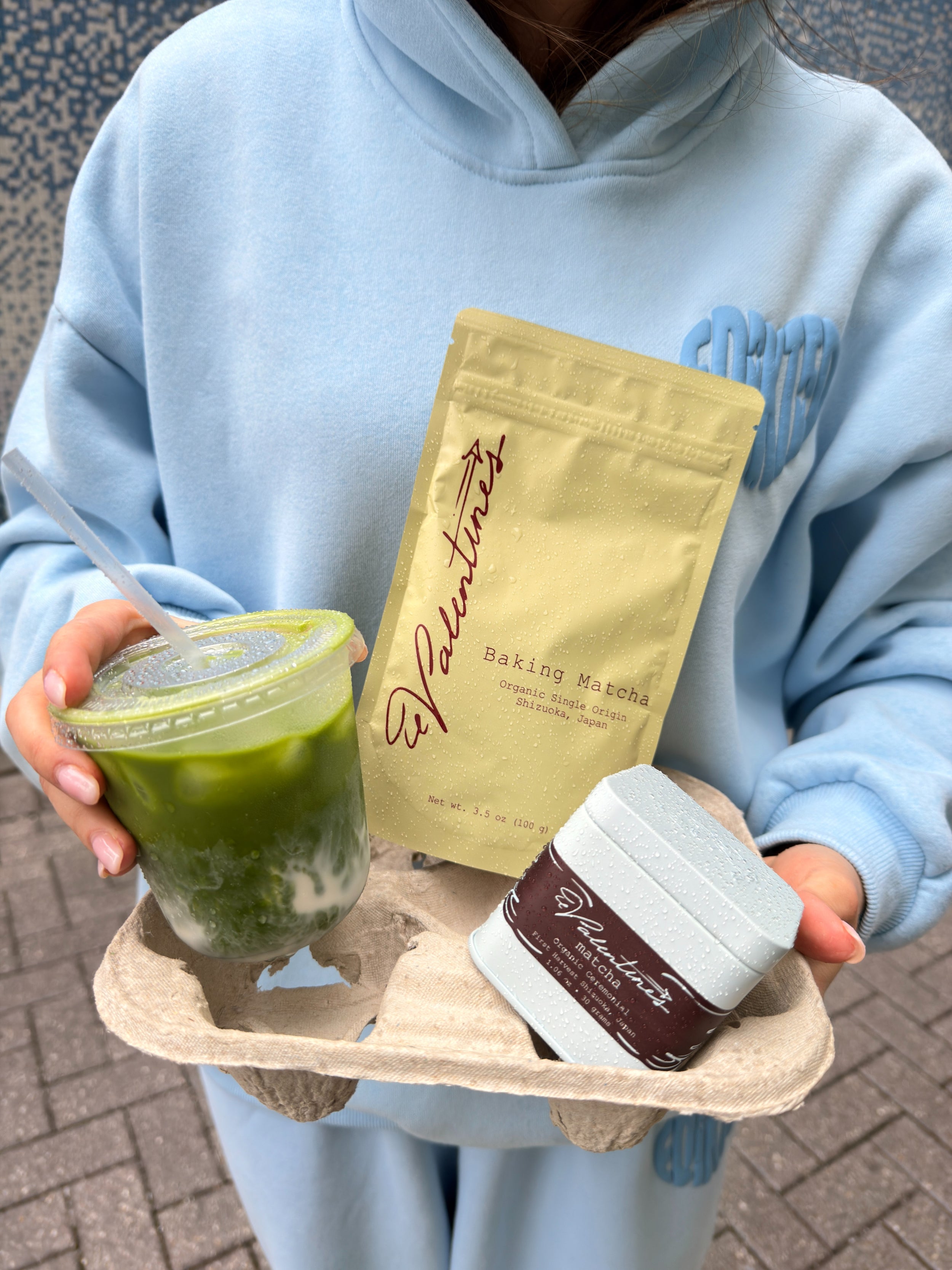 person holding iced matcha latte with baking matcha pouch and blue ceremonial matcha tin in cardboard drink tray