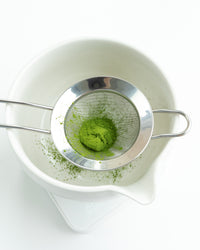 Silver mesh strainer with green matcha powder over a white bowl on a white background