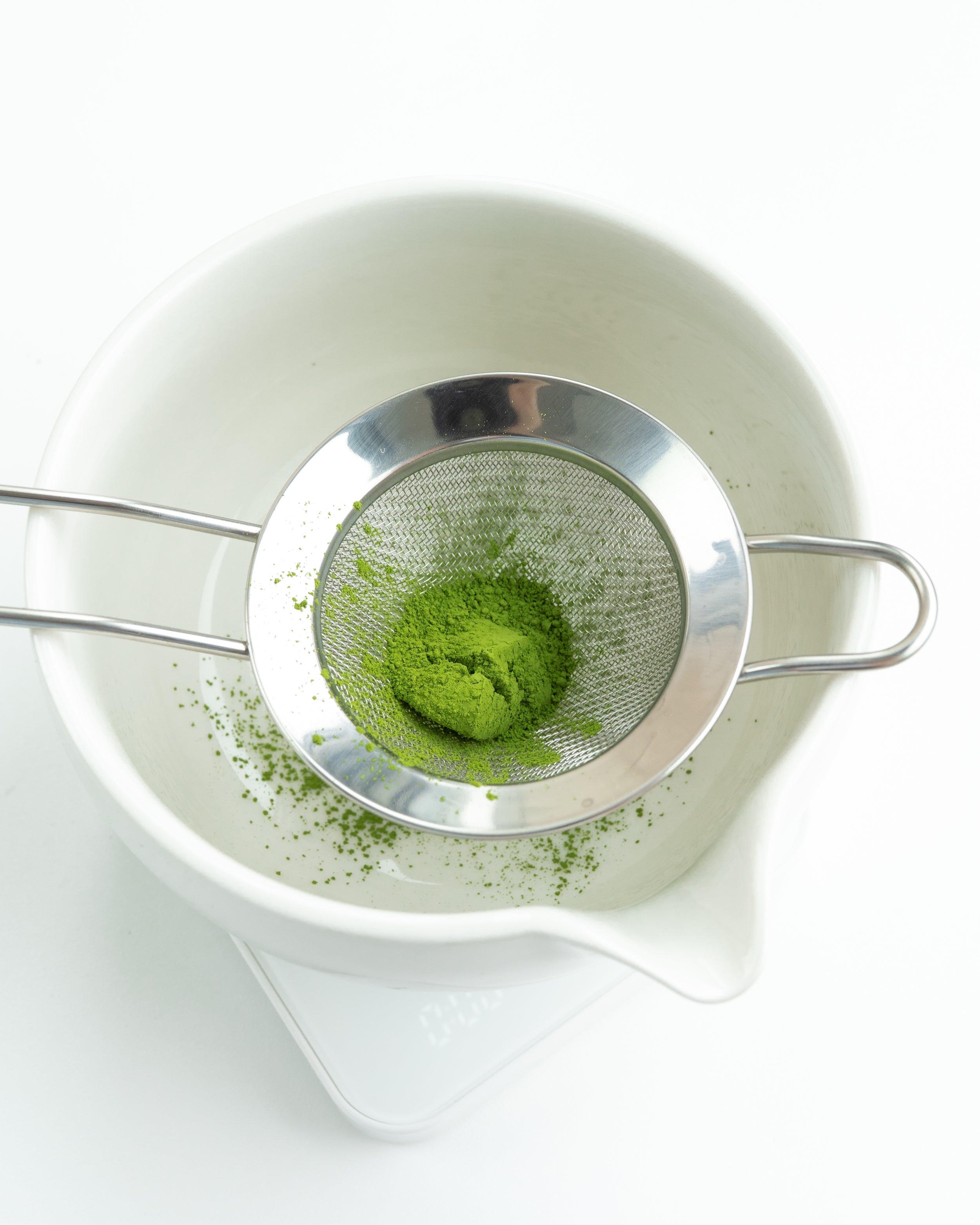 Silver mesh strainer with green matcha powder over a white bowl on a white background