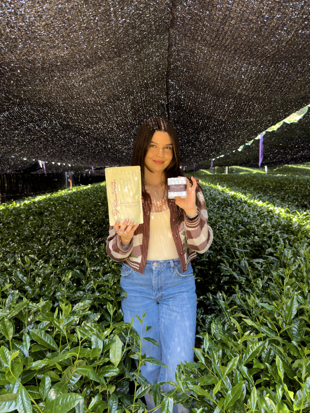 Valentina holding a tin and a pouch of matcha in a tea field under shade cloth