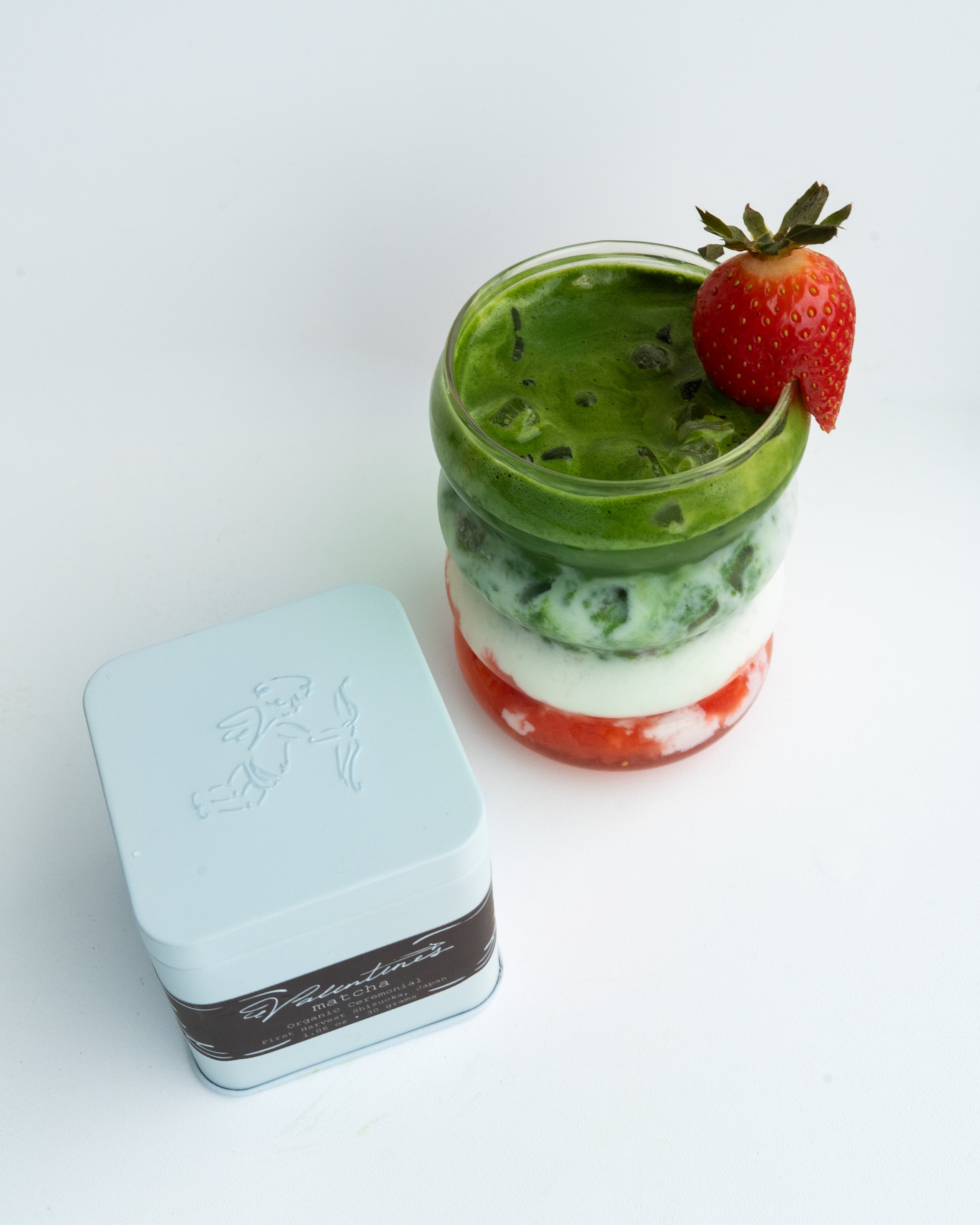 Stacked layered drink with a strawberry on top next to a light blue tin on a white background