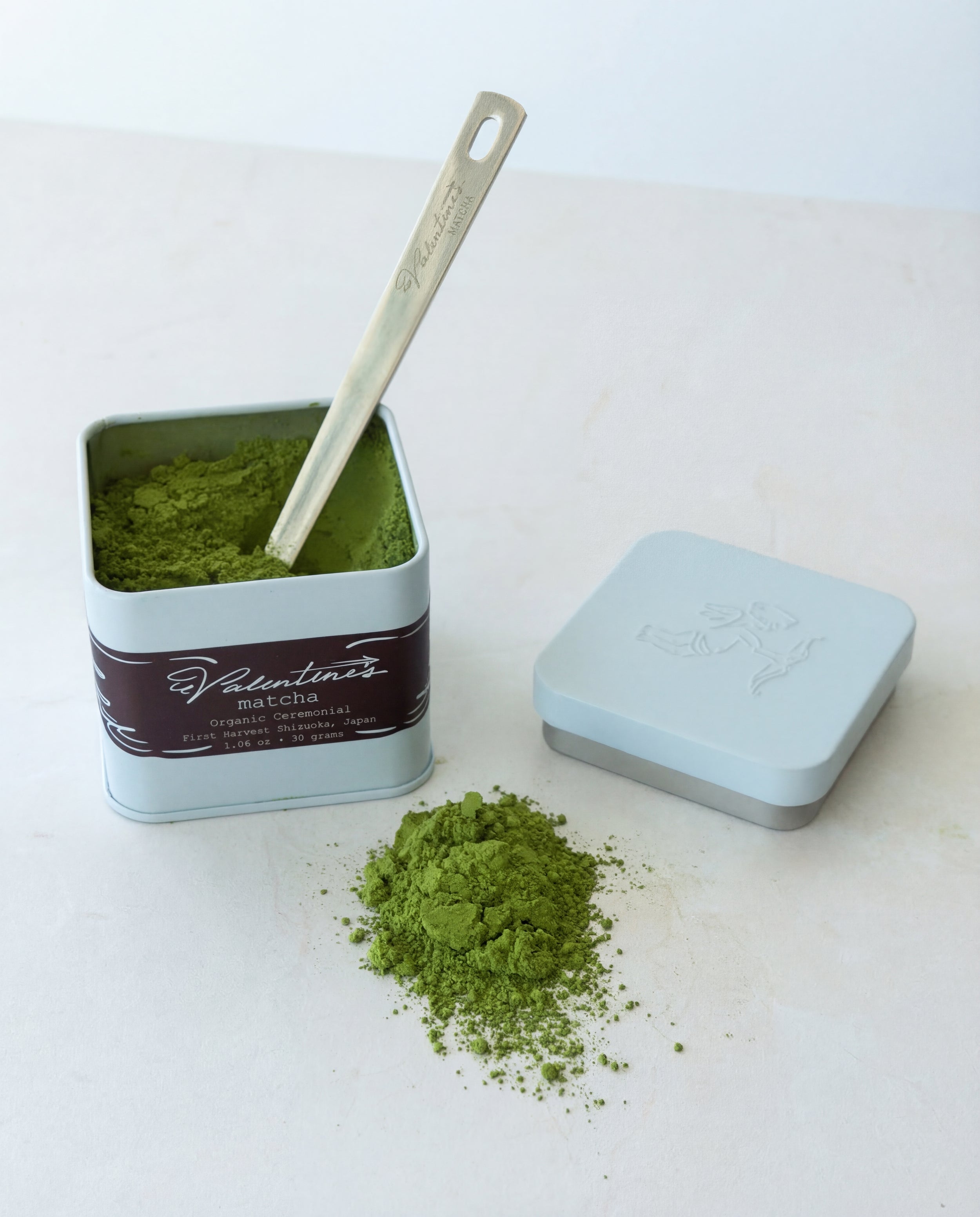 Matcha green tea powder in a blue tin with a scoop on a white surface and matcha power on the white surface